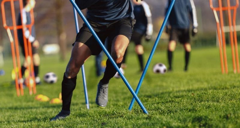 African-American soccer player on training drill. Legs of footballer running on grass practice field. Obstacle course in soccer football. Player running in sports cleats
