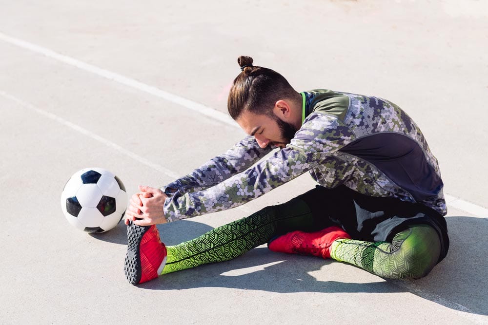 football player stretching legs sitting on the floor next to his football ball in a concrete soccer court, concept of healthy lifestyle and urban sport in the city, copy space for text