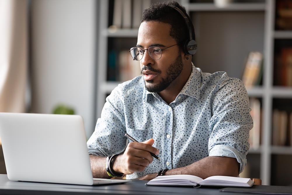Focused young african businessman wear headphones study online watching webinar podcast on laptop listening learning education course conference calling make notes sit at work desk, elearning concept