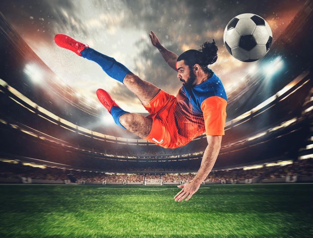 Soccer striker with orange and blue uniform hits the ball with an acrobatic kick in the air at the stadium