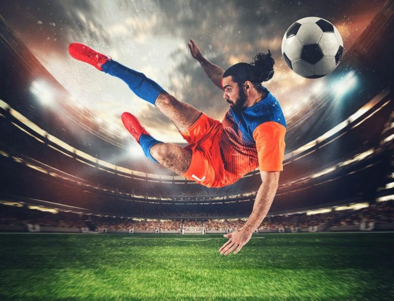 Soccer striker with orange and blue uniform hits the ball with an acrobatic kick in the air at the stadium